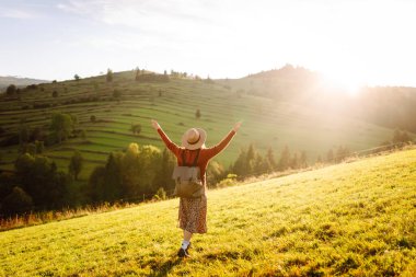 Parlak yeşil vadiyi gezen sırt çantalı güzel bir turist. Şapkalı genç bir kadın günbatımının ve doğanın tadını çıkarıyor. Seyahat, doğa ve özgürlük kavramı. Etkin yaşam biçimi.