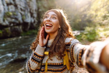 Neşeli genç bir kadın telefonuyla dağ nehrinin yanında selfie çekiyor. Sırt çantalı güzel bayan gezgin doğada günbatımının tadını çıkarıyor. Bloglama, yürüyüş kavramı.