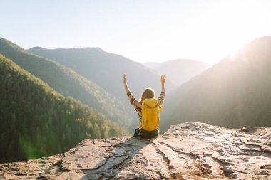 Genç bir kadın sırtını kayalık bir uçurumun kenarına dayamış dağ manzarasına bakıyor. Sırt çantalı kadın gezgin gün batımının tadını çıkarıyor, özgür hissediyor. Doğa ve keyif kavramı.