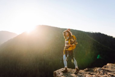 Genç bir kadın sırtını kayalık bir uçurumun kenarına dayamış dağ manzarasına bakıyor. Sırt çantalı kadın gezgin gün batımının tadını çıkarıyor, özgür hissediyor. Doğa ve keyif kavramı.