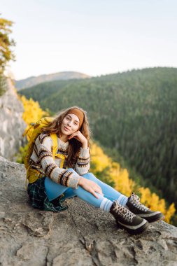 Genç bir kadın sırtını kayalık bir uçurumun kenarına dayamış dağ manzarasına bakıyor. Sırt çantalı kadın gezgin gün batımının tadını çıkarıyor, özgür hissediyor. Doğa ve keyif kavramı.