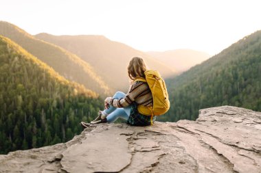 Genç bir kadın sırtını kayalık bir uçurumun kenarına dayamış dağ manzarasına bakıyor. Sırt çantalı kadın gezgin gün batımının tadını çıkarıyor, özgür hissediyor. Doğa ve keyif kavramı.