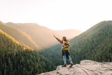 Genç bir kadın sırtını kayalık bir uçurumun kenarına dayamış dağ manzarasına bakıyor. Sırt çantalı kadın gezgin gün batımının tadını çıkarıyor, özgür hissediyor. Doğa ve keyif kavramı.