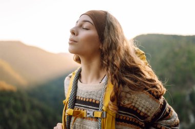 Genç bir kadın sırtını kayalık bir uçurumun kenarına dayamış dağ manzarasına bakıyor. Sırt çantalı kadın gezgin gün batımının tadını çıkarıyor, özgür hissediyor. Doğa ve keyif kavramı.