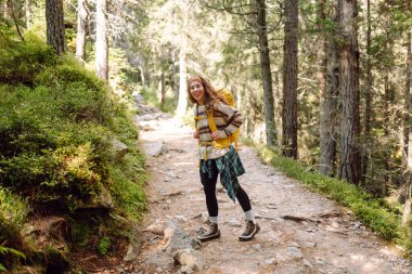 Sırt çantalı genç bayan yürüyüşçü yemyeşil ormanda kayalık yolda yürüyor. Güneşli bir günde doğa ile eğlenen mutlu kadın yürüyüşçü. Macera, eğlence konsepti. Etkin yaşam biçimi.