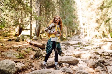 Sırt çantalı genç bayan yürüyüşçü yemyeşil ormanda kayalık yolda yürüyor. Güneşli bir günde doğa ile eğlenen mutlu kadın yürüyüşçü. Macera, eğlence konsepti. Etkin yaşam biçimi.
