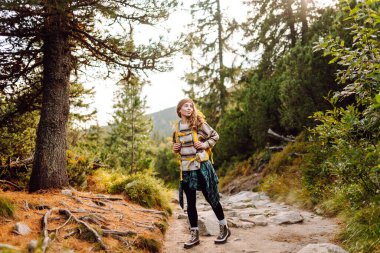 Sırt çantalı genç bayan yürüyüşçü yemyeşil ormanda kayalık yolda yürüyor. Güneşli bir günde doğa ile eğlenen mutlu kadın yürüyüşçü. Macera, eğlence konsepti. Etkin yaşam biçimi.