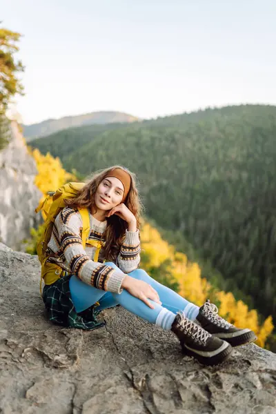 Genç bir kadın sırtını kayalık bir uçurumun kenarına dayamış dağ manzarasına bakıyor. Sırt çantalı kadın gezgin gün batımının tadını çıkarıyor, özgür hissediyor. Doğa ve keyif kavramı.