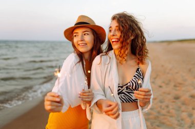 Havai fişekli güzel kız arkadaşlar gün batımı kumsalındaki anın tadını çıkarıyorlar. İki genç arkadaş birlikte vakit geçiriyorlar, dışarıda ışıkların tadını çıkarıyorlar. Kutlama kavramı, özgürlük..