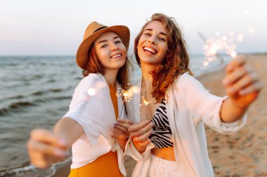 Havai fişekli güzel kız arkadaşlar gün batımı kumsalındaki anın tadını çıkarıyorlar. İki genç arkadaş birlikte vakit geçiriyorlar, dışarıda ışıkların tadını çıkarıyorlar. Kutlama kavramı, özgürlük..