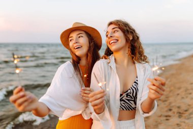 Havai fişekli güzel kız arkadaşlar gün batımı kumsalındaki anın tadını çıkarıyorlar. İki genç arkadaş birlikte vakit geçiriyorlar, dışarıda ışıkların tadını çıkarıyorlar. Kutlama kavramı, özgürlük..