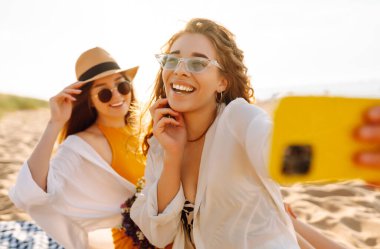 Sahilde eğlenen ve gün batımında selfie çeken iki genç kadın. Güzel kızlar birlikte zaman geçirir ve kumlu bir sahilde yürürler. Arkadaşlık kavramı. Selfie zamanı..