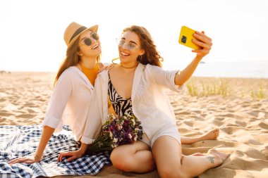 Sahilde eğlenen ve gün batımında selfie çeken iki genç kadın. Güzel kızlar birlikte zaman geçirir ve kumlu bir sahilde yürürler. Arkadaşlık kavramı. Selfie zamanı..