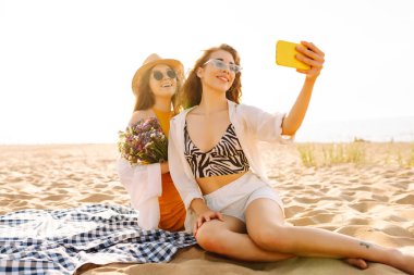 Sahilde eğlenen ve gün batımında selfie çeken iki genç kadın. Güzel kızlar birlikte zaman geçirir ve kumlu bir sahilde yürürler. Arkadaşlık kavramı. Selfie zamanı..