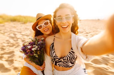 Sahilde eğlenen ve gün batımında selfie çeken iki genç kadın. Güzel kızlar birlikte zaman geçirir ve kumlu bir sahilde yürürler. Arkadaşlık kavramı. Selfie zamanı..