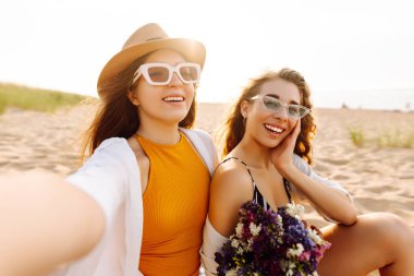 Sahilde eğlenen ve gün batımında selfie çeken iki genç kadın. Güzel kızlar birlikte zaman geçirir ve kumlu bir sahilde yürürler. Arkadaşlık kavramı. Selfie zamanı..