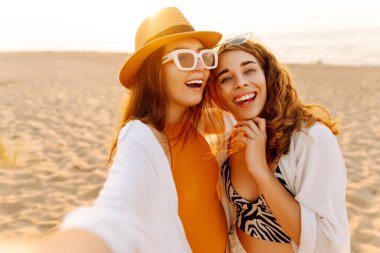 Sahilde eğlenen ve gün batımında selfie çeken iki genç kadın. Güzel kızlar birlikte zaman geçirir ve kumlu bir sahilde yürürler. Arkadaşlık kavramı. Selfie zamanı..