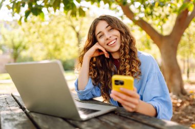 Telefonlu ve bilgisayarlı genç bir kadın güneşli bir parkta çalışıyor. Güzel bayan serbest çalışan hava durumunun ve telefonunda mesajlaşmanın tadını çıkarıyor. Uzak çalışma konsepti.