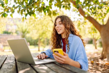 Telefonlu ve bilgisayarlı genç bir kadın güneşli bir parkta çalışıyor. Güzel bayan serbest çalışan hava durumunun ve telefonunda mesajlaşmanın tadını çıkarıyor. Uzak çalışma konsepti.