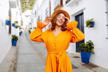 Dağlarda küçük bir kasabada gezen şapkalı ve parlak elbiseli bir kadın turist. Yürüyüşten, havadan ve mimariden hoşlanan güzel bir kadın. Seyahat, yürüyüş konsepti.