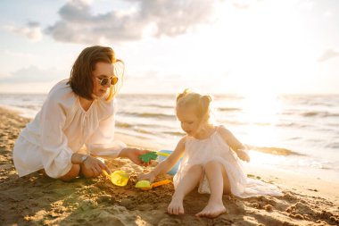 Genç anne ve kızı kumsalda kum inşa ederek eğleniyorlar. Güzel bir kadın ve bir çocuk dışarıda birlikte vakit geçiriyorlar. Çocukluk kavramı, annelik, eğlence..