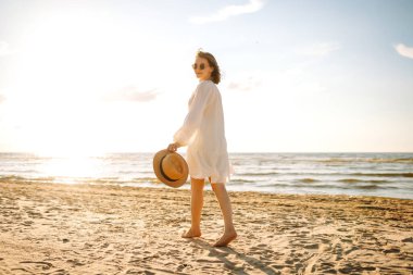 Şapkalı ve güneş gözlüklü güzel bir kadın kumlu sahilde yürüyor ve saçlarıyla rüzgârı yakalıyor. Genç bir kadın güneşli havanın tadını çıkarıyor, deniz manzaralı özgürlüğün tadını çıkarıyor. Keyif ve özgürlük kavramı.