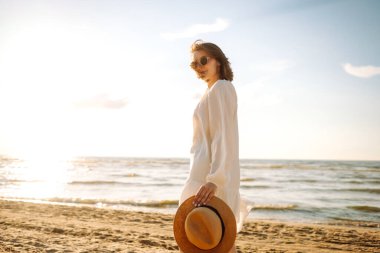 Şapkalı ve güneş gözlüklü güzel bir kadın kumlu sahilde yürüyor ve saçlarıyla rüzgârı yakalıyor. Genç bir kadın güneşli havanın tadını çıkarıyor, deniz manzaralı özgürlüğün tadını çıkarıyor. Keyif ve özgürlük kavramı.