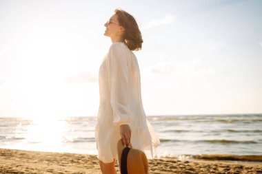 Şapkalı ve güneş gözlüklü güzel bir kadın kumlu sahilde yürüyor ve saçlarıyla rüzgârı yakalıyor. Genç bir kadın güneşli havanın tadını çıkarıyor, deniz manzaralı özgürlüğün tadını çıkarıyor. Keyif ve özgürlük kavramı.