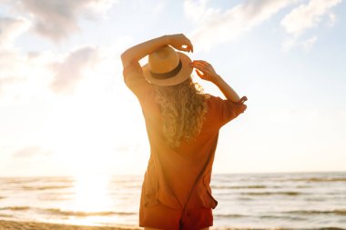 Kıvırcık saçlı genç bir kadının kıyı şeridinde rahat hisseder. Genç bir kadın gün batımında sahilde yürür. Rahatlama, keyif, özgürlük kavramı.