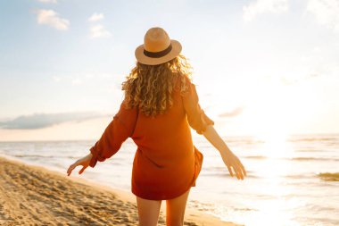 Kıvırcık saçlı genç bir kadının kıyı şeridinde rahat hisseder. Genç bir kadın gün batımında sahilde yürür. Rahatlama, keyif, özgürlük kavramı.