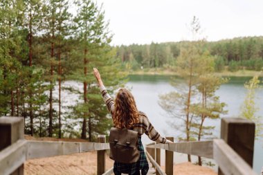 Sırt çantalı genç bir kadın ormandaki ahşap patikalarda yürüyüş yapıyor. Güzel bayan gezgin özgürlüğü hissediyor ve göl manzarasının tadını çıkarıyor. Yürüyüş, doğa konsepti..