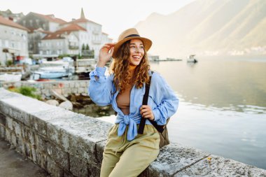 Hasır şapkalı bir kadın gezgin, pitoresk bir limanda yürüyor. Genç kadın deniz manzarası ve güneşli havanın tadını çıkarıyor. Seyahat, özgürlük konsepti. Etkin yaşam biçimi.