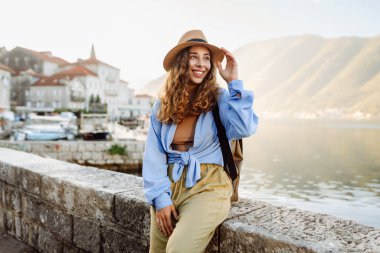 Hasır şapkalı bir kadın gezgin, pitoresk bir limanda yürüyor. Genç kadın deniz manzarası ve güneşli havanın tadını çıkarıyor. Seyahat, özgürlük konsepti. Etkin yaşam biçimi.