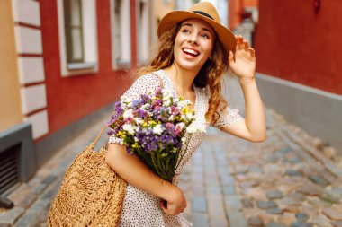 Güneşli Avrupa caddesinde yürüyen şapkalı ve buketi olan güzel bir kadın. Genç turist dışarıda eğleniyor ve eğleniyor. Yürüyen konsept, seyahat.