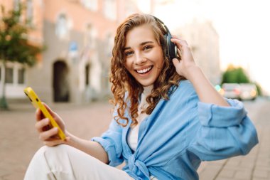 Dışarıda kablosuz kulaklık ve telefonla müzik dinleyen mutlu bir kadının portresi. Telefonlu ve kulaklıklı genç bir kadın güneşli şehir caddesinde yürüyor. Yürüyüş kavramı, rahatlama..