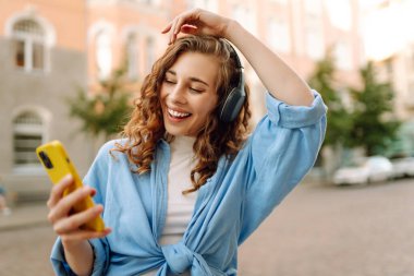 Dışarıda kablosuz kulaklık ve telefonla müzik dinleyen mutlu bir kadının portresi. Telefonlu ve kulaklıklı genç bir kadın güneşli şehir caddesinde yürüyor. Yürüyüş kavramı, rahatlama..