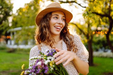 Güzel bir buket kır çiçeğiyle genç bir turist bir yaz parkında eğleniyor. Hasır şapkalı güzel bir kadın güneşli havanın tadını çıkarır. Güzellik, tatil, rahatlama kavramı..