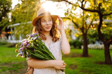 Güzel bir buket kır çiçeğiyle genç bir turist bir yaz parkında eğleniyor. Hasır şapkalı güzel bir kadın güneşli havanın tadını çıkarır. Güzellik, tatil, rahatlama kavramı..
