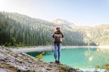 Sırt çantalı genç bir bayan yürüyüşçü gün doğumunda bir dağ gölü manzarasının keyfini çıkarıyor. Şapkalı güzel bayan yürüyüşçü dağ manzaralı özgürlüğü hissediyor. Macera, doğa ve yürüyüş konsepti.