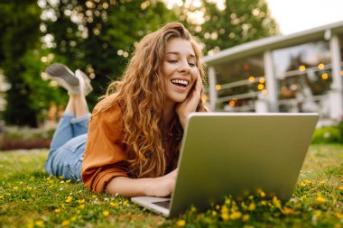 Bilgisayarlı genç bir kadın parkta yeşil çimlerde çalışıyor. Dizüstü bilgisayarı olan serbest çalışan bir kadın parkta güneşli havanın tadını çıkarıyor. Uzaktan çalışma konsepti, serbest yazar. Yaşam biçimi.