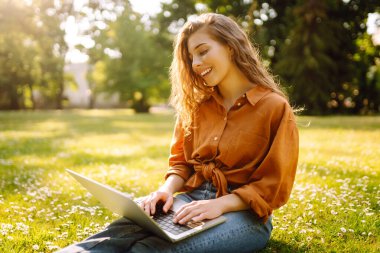 Bilgisayarlı genç bir kadın parkta yeşil çimlerde çalışıyor. Dizüstü bilgisayarı olan serbest çalışan bir kadın parkta güneşli havanın tadını çıkarıyor. Uzaktan çalışma konsepti, serbest yazar. Yaşam biçimi.