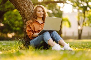 Bilgisayarlı genç bir kadın parkta yeşil çimlerde çalışıyor. Dizüstü bilgisayarı olan serbest çalışan bir kadın parkta güneşli havanın tadını çıkarıyor. Uzaktan çalışma konsepti, serbest yazar. Yaşam biçimi.