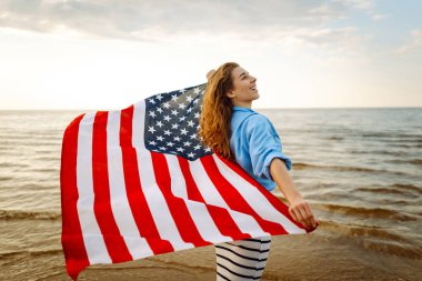 Amerikan bayrağıyla gün batımında kumlu sahilde vızıldayan mutlu kadın portresi. Özgürlüğün tadını çıkaran genç bir kadın. Bağımsızlık kavramı.