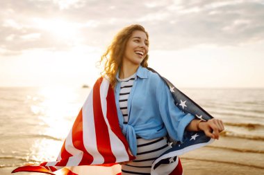 Amerikan bayrağıyla gün batımında kumlu sahilde vızıldayan mutlu kadın portresi. Özgürlüğün tadını çıkaran genç bir kadın. Bağımsızlık kavramı.