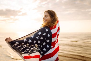 Amerikan bayrağıyla gün batımında kumlu sahilde vızıldayan mutlu kadın portresi. Özgürlüğün tadını çıkaran genç bir kadın. Bağımsızlık kavramı.
