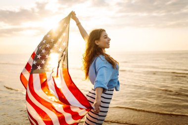 Amerikan bayrağıyla gün batımında kumlu sahilde vızıldayan mutlu kadın portresi. Özgürlüğün tadını çıkaran genç bir kadın. Bağımsızlık kavramı.