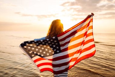 Amerikan bayrağıyla gün batımında kumlu sahilde vızıldayan mutlu kadın portresi. Özgürlüğün tadını çıkaran genç bir kadın. Bağımsızlık kavramı.