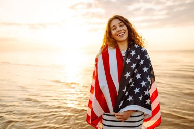 Amerikan bayrağıyla gün batımında kumlu sahilde vızıldayan mutlu kadın portresi. Özgürlüğün tadını çıkaran genç bir kadın. Bağımsızlık kavramı.