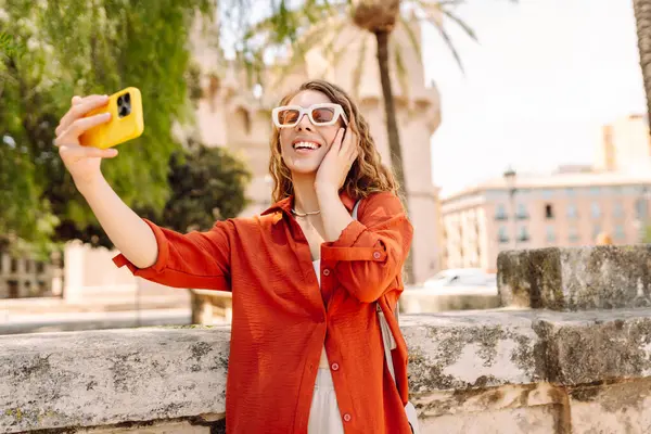 Şık kıyafetler içinde gülümseyen bir turist ve şehrin güneşli caddelerinde telefonla yürüyor. Kadın dışarıda cep telefonuyla selfie ve blog çekiyor. Bloglama, seyahat kavramı.