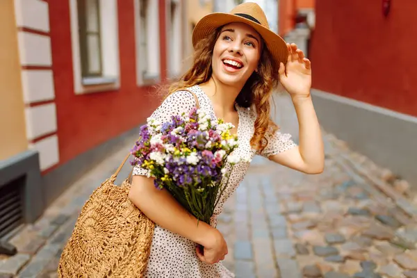Güneşli Avrupa caddesinde yürüyen şapkalı ve buketi olan güzel bir kadın. Genç turist dışarıda eğleniyor ve eğleniyor. Yürüyen konsept, seyahat.